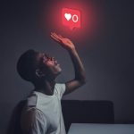 A man sits indoors reaching towards a glowing social media notification icon.