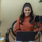 Young woman in a sweater podcasting indoors with a microphone and laptop, smiling confidently.